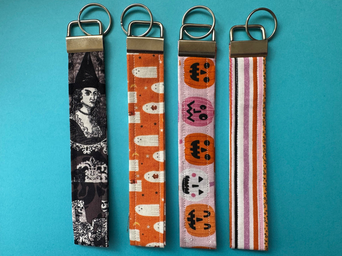 Four Halloween-themed fabric keychains laid flat on a bright blue background. The keychains feature different spooky patterns: a black and white witch illustration, white ghosts on orange, jack-o'-lanterns on pink, and orange and white stripes.